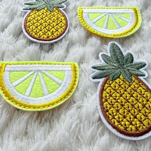 May include: Four embroidered fruit patches: two pineapples and two lemon slices. The pineapples have green tops and yellow bodies, while the lemon slices are yellow and white. They are displayed on a white, fluffy background.
