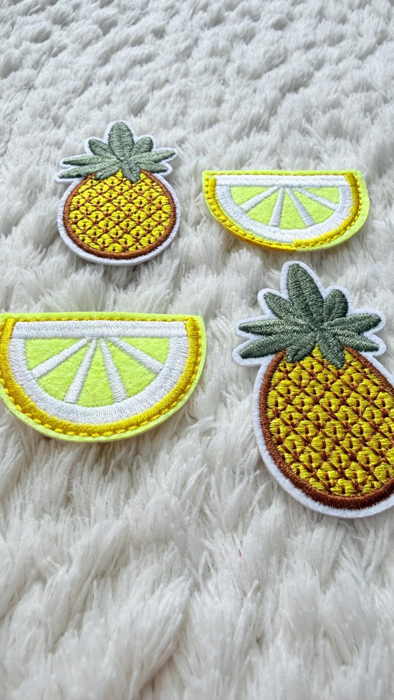 May include: Four embroidered fruit patches: two pineapples and two lemon slices. The pineapples have green tops and yellow bodies, while the lemon slices are yellow and white. They are displayed on a white, fluffy background.