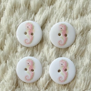 Puede incluir: Cuatro botones blancos con un diseño de caballito de mar rosa. Los caballitos de mar tienen una apariencia delicada, similar a la acuarela, con detalles rosas y verdes. Cada botón tiene dos agujeros para coser.