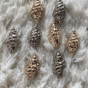 May include: Nine seashell-shaped charms in gold and silver hues. The charms have a textured surface and are displayed on a white, fluffy background. These decorative charms are suitable for jewellery making or craft projects.