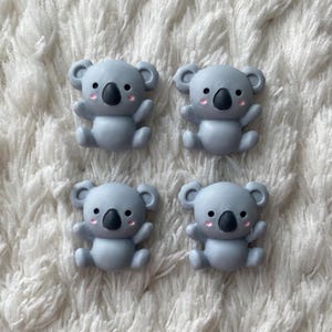 Peut inclure: Quatre figurines de koala gris avec des yeux noirs et des joues roses. Les koalas sont assis les bras tendus.