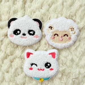 Può includere: Tre toppe ricamate con facce di animali: un panda con orecchie nere, una pecora con la faccia beige e un gatto con orecchie rosse e collare blu. Le toppe sono bianche con dettagli neri e rosa.