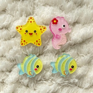 Può includere: Quattro ornamenti di creature marine in plastica colorata. Una stella marina gialla, un cavalluccio marino rosa e due pesci blu e verdi.