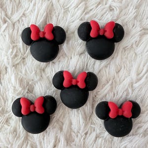 Könnte beinhalten: Fünf schwarze Minnie-Maus-förmige Verzierungen mit leuchtend roten Schleifen. Die Verzierungen sind auf einer weißen, strukturierten Oberfläche angeordnet. Diese könnten zum Basteln oder Dekorieren verwendet werden.