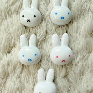 May include: Five white ceramic bunny figurines with black or colored facial features. The bunnies have a minimalist design with small black or colored dots for eyes and an X-shaped mouth. The figurines are arranged on a fluffy, white surface.