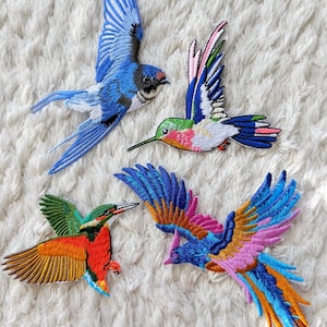 Peut inclure: Quatre écussons brodés représentant des oiseaux avec un travail de fil détaillé. Un oiseau bleu à longue queue, un colibri aux ailes roses, vertes et bleues, un oiseau vert et orange à long bec, et un oiseau multicolore aux ailes bleues et roses.