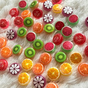 Op de afbeelding: Een verzameling kleurrijke hars cabochons met fruit thema's. De cabochons tonen gedetailleerde ontwerpen van sinaasappels, kiwi's, watermeloenen en ander fruit. Ze zijn verspreid op een zacht, wit, pluizig oppervlak.