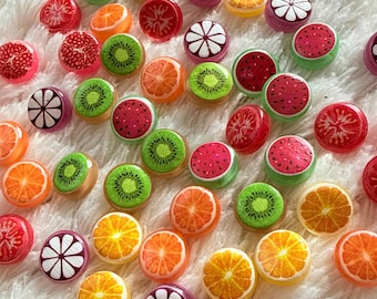 May include: A collection of colorful, fruit-themed resin cabochons. The cabochons feature detailed designs of oranges, kiwis, watermelons, and other fruits. They are scattered on a soft, white, fluffy surface.