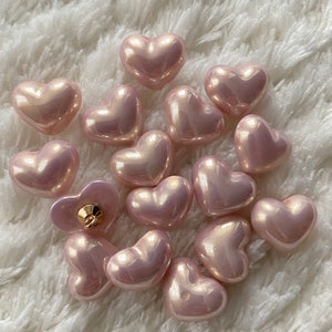 May include: A collection of 15 pink heart-shaped buttons with a pearlescent finish. The buttons are arranged in a cluster on a white fluffy surface.