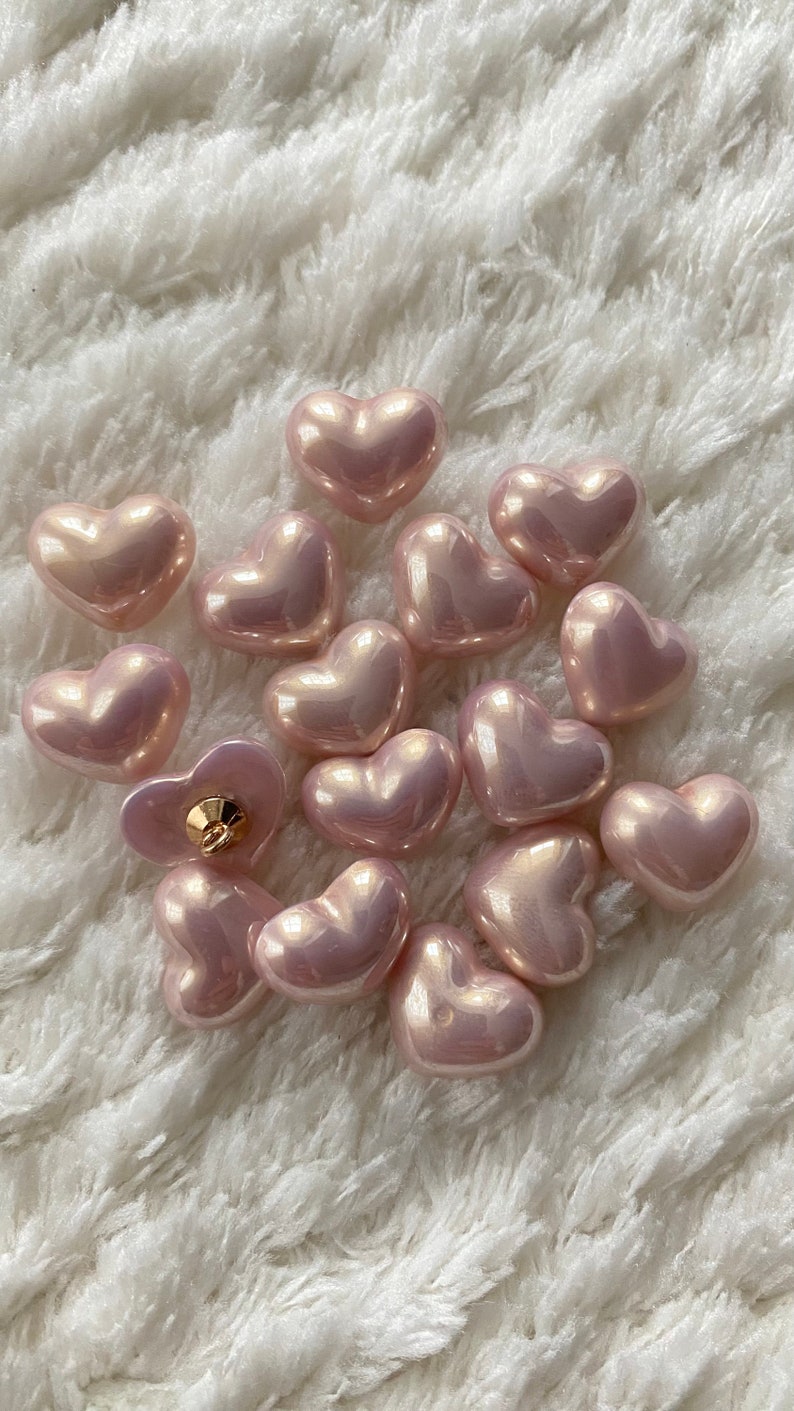 May include: A collection of 15 pink heart-shaped buttons with a pearlescent finish. The buttons are arranged in a cluster on a white fluffy surface.