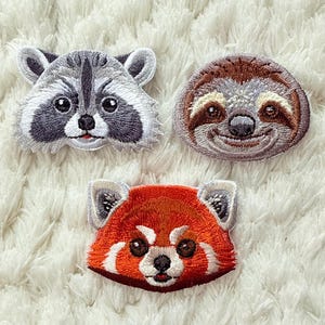 May include: Embroidered patches featuring animal faces: a raccoon, a sloth, and a red panda. The raccoon patch is gray and white, the sloth is brown, and the red panda is red and white. Each patch has detailed stitching and a friendly expression.
