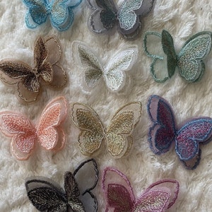 Può includere: Assortimento di dieci applicazioni a forma di farfalla in vari colori, tra cui blu, verde, rosa, oro e argento. Le applicazioni sono realizzate in tessuto trasparente con dettagli ricamati.