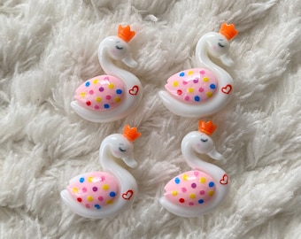 May include: Four white swan-shaped resin embellishments with pink, yellow, blue, and red polka dots, orange crowns, and a small pink heart on each.