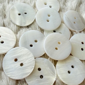 May include: A collection of round, white, iridescent buttons, each featuring two centered holes. The buttons have a smooth, polished surface with a subtle, natural pearlescent sheen, displaying varying textures and light reflections across their circular forms.