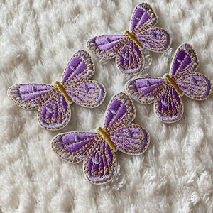 May include: Four purple and gold embroidered butterfly patches on a white fluffy surface.