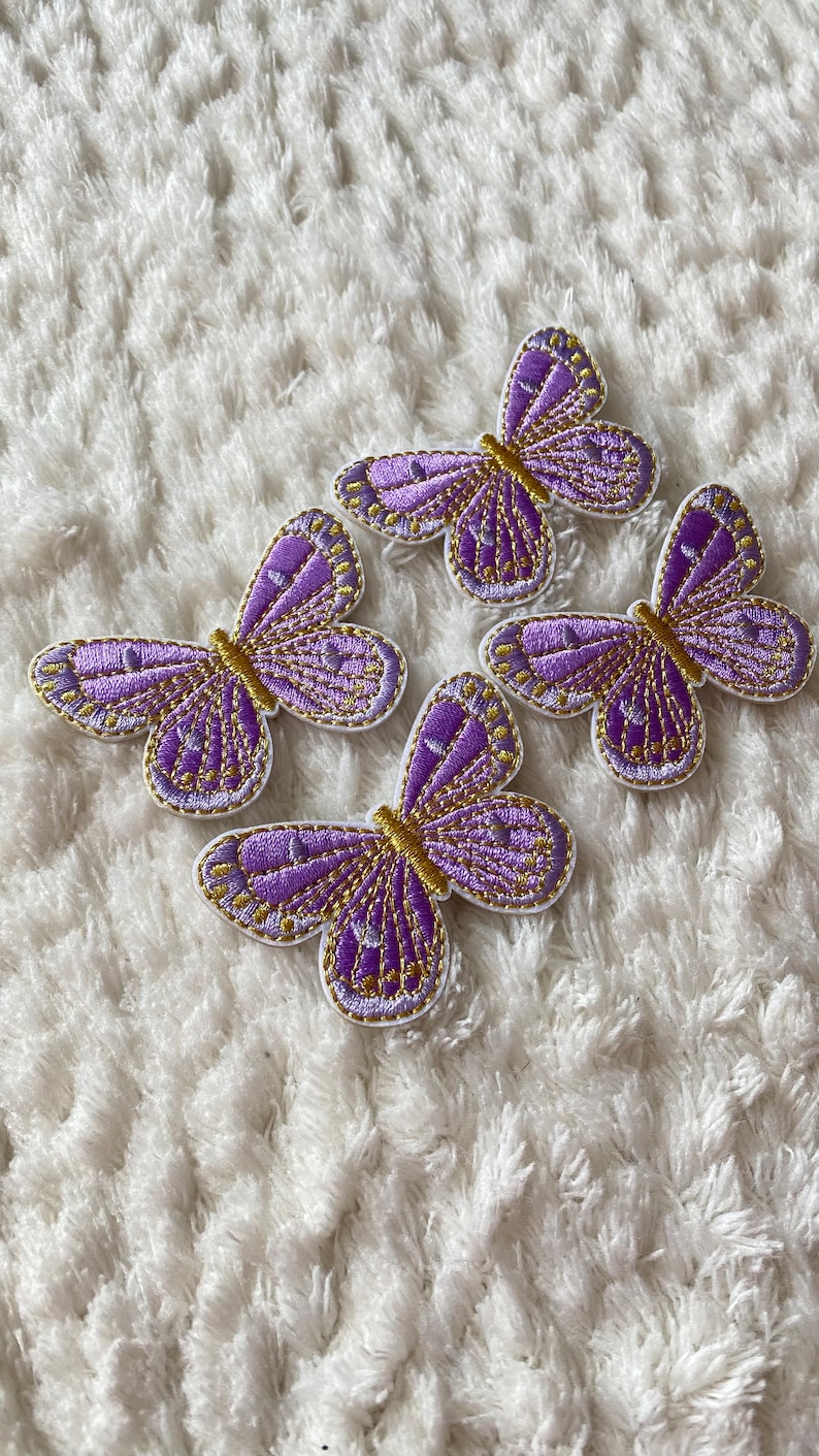 May include: Four purple and gold embroidered butterfly patches on a white fluffy surface.