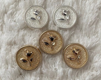 May include: Four round buttons with a raised rose design. Two buttons are gold with a white rose, and two buttons are silver with a white rose.