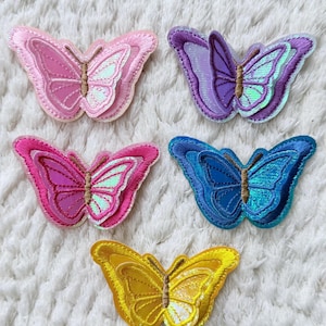May include: Five embroidered butterfly patches in shades of pink, purple, blue, and yellow. Each butterfly has detailed wing patterns and a brown body. The patches are arranged on a white, textured surface.