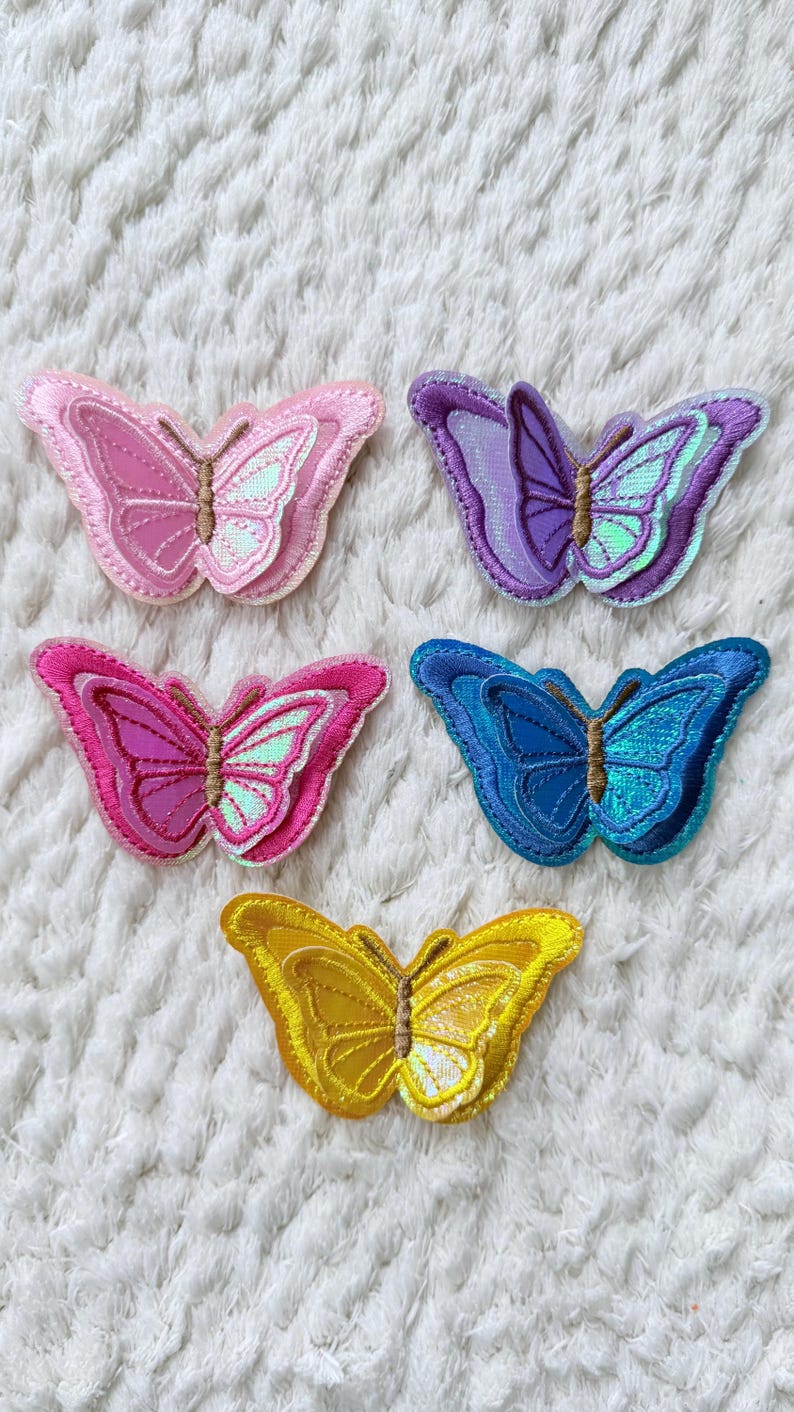 May include: Five embroidered butterfly patches in shades of pink, purple, blue, and yellow. Each butterfly has detailed wing patterns and a brown body. The patches are arranged on a white, textured surface.