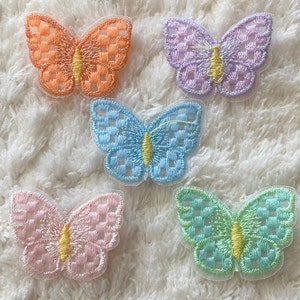 Könnte beinhalten: Fünf gestickte Schmetterlings-Patches in verschiedenen Pastellfarben, darunter hellblau, rosa, grün, lila und orange. Jeder Schmetterling hat ein kariertes Muster und eine gelbe Mitte.
