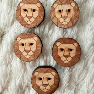 Puede incluir: Cinco botones de madera con caras de león. Cada botón presenta un diseño geométrico de una cara de león con una melena y cara marrón claro, y un contorno marrón oscuro. Los botones son redondos y tienen dos pequeños agujeros para coser.