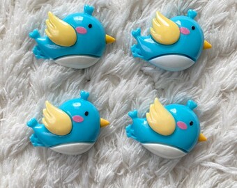 May include: Four blue bird-shaped embellishments with yellow wings, pink cheeks, and yellow beaks. The birds are on a white, textured surface. These decorative elements are suitable for crafts and DIY projects.
