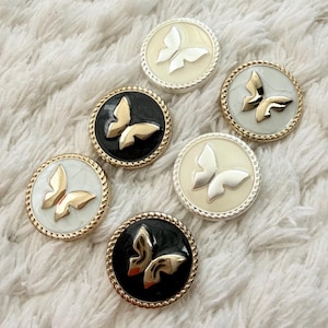 Peut inclure: Ensemble de sept boutons décoratifs avec des motifs de papillons. Chaque bouton présente un papillon doré sur un fond noir, blanc ou crème, bordé d'un liseré doré ou argenté en forme de corde. Les boutons sont ronds et brillants.