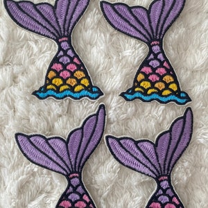 May include: Four purple and multi-colored mermaid tail iron-on patches. Each patch features a purple tail with a pink, yellow, and blue scale pattern.