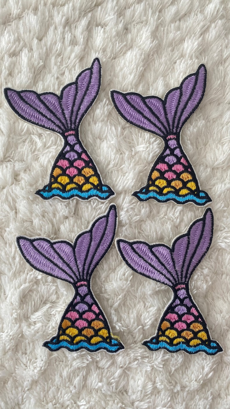 May include: Four purple and multi-colored mermaid tail iron-on patches. Each patch features a purple tail with a pink, yellow, and blue scale pattern.