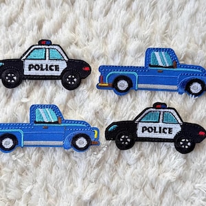 Puede incluir: Cuatro parches bordados con vehículos policiales. Dos son coches de policía en blanco y negro con el texto "POLICE", y dos son camionetas azules. Todos tienen costuras detalladas y están sobre un fondo blanco texturizado.
