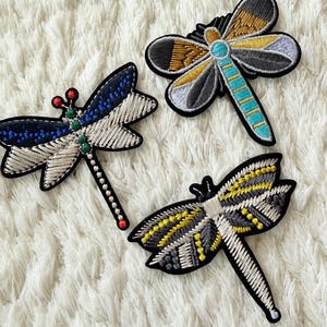May include: Three embroidered dragonfly patches featuring detailed stitching. One patch has blue and silver wings with red and green accents. Another has gold, grey, and turquoise wings. The third features yellow, grey, and white wing patterns.