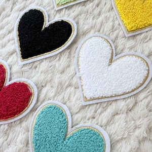 Puede incluir: Un conjunto de parches en forma de corazón en varios colores, incluyendo verde, amarillo, negro, blanco, rojo y turquesa. Cada parche tiene una apariencia texturizada y difusa, con un borde dorado y un borde blanco. Los parches están dispuestos sobre una superficie blanca texturizada.