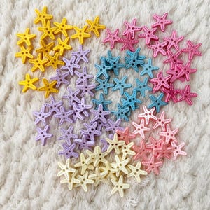 Op de afbeelding: Een verzameling kleurrijke knopen in de vorm van zeesterren in geel, lavendel, lichtblauw, roze en crème. De knopen liggen op een witte, getextureerde ondergrond. Perfect voor naai- en knutselprojecten.