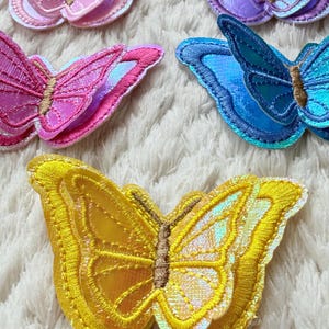 May include: Embroidered butterfly patches in various colors: pink, purple, blue, and yellow. Each patch features detailed stitching and iridescent accents, creating a shimmering effect. The butterflies have brown centers and are arranged on a white, textured surface.