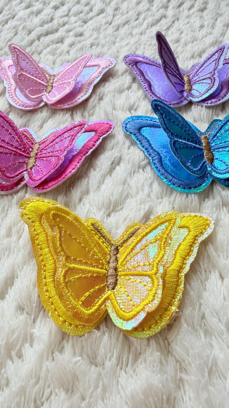 May include: Embroidered butterfly patches in various colors: pink, purple, blue, and yellow. Each patch features detailed stitching and iridescent accents, creating a shimmering effect. The butterflies have brown centers and are arranged on a white, textured surface.