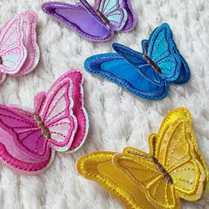 May include: A collection of colorful butterfly patches in various shades of pink, purple, blue, and yellow. Each patch features detailed stitching and iridescent accents, creating a shimmering effect. The patches are arranged on a white, textured surface.