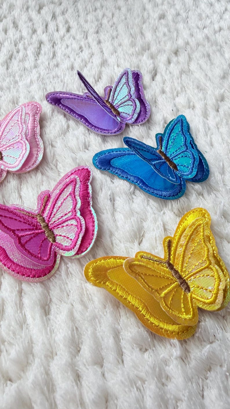 May include: A collection of colorful butterfly patches in various shades of pink, purple, blue, and yellow. Each patch features detailed stitching and iridescent accents, creating a shimmering effect. The patches are arranged on a white, textured surface.