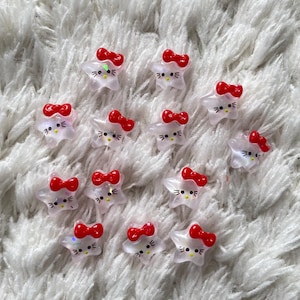 Puede incluir: Adornos transparentes en forma de estrella con lazos rojos y caras de gato. Estas pequeñas piezas decorativas están dispersas sobre una superficie blanca y esponjosa. Los adornos presentan un lazo rojo y una cara de gato con ojos y bigotes negros.