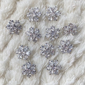 Op de afbeelding: Twaalf zilveren bloemvormige versieringen met heldere strass steentjes. Elke versiering heeft een cluster van peervormige strass steentjes.