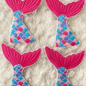 May include: Four pink mermaid tail patches with blue, pink, and peach scales. The patches are embroidered and have a white outline.