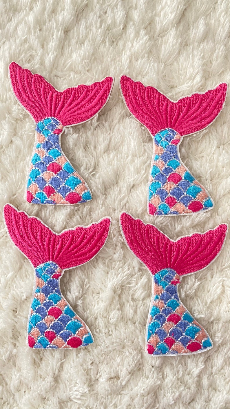 May include: Four pink mermaid tail patches with blue, pink, and peach scales. The patches are embroidered and have a white outline.