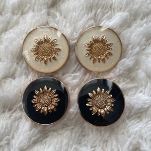 Può includere: Quattro bottoni rotondi con un designo a girasole dorato. Due bottoni hanno uno sfondo bianco con glitter dorato e due bottoni hanno uno sfondo nero.