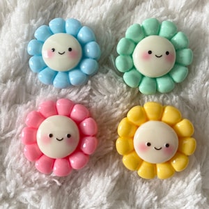 Può includere: Quattro ciondoli in resina colorati a forma di fiore con facce sorridenti. I ciondoli sono azzurro, verde, rosa e giallo.