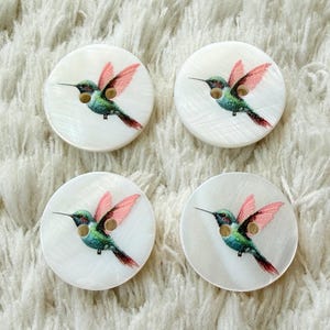 Könnte beinhalten: Vier runde, weiße, schimmernde Perlmuttknöpfe, die jeweils mit einem aufgedruckten Kolibri mit grünem Körper und rosa Flügeln versehen sind. Jeder Knopf hat zwei mittige Löcher zur Befestigung und eine glatte, polierte Oberfläche.