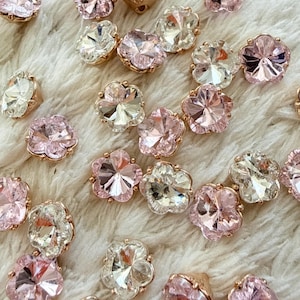 May include: A collection of small, flower-shaped buttons featuring faceted crystals in shades of pale pink and clear white. Each button is set in a metallic base, showcasing a multi-petaled design that reflects light through its polished surfaces.