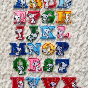May include: Colorful embroidered alphabet letters featuring Snoopy. Each letter is a different color, with Snoopy in various poses. The letters are arranged on a white, textured surface. A fun and playful design for crafts or personalization.