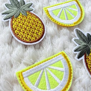 May include: Embroidered patches of a pineapple and lemon slices. The pineapple patch is yellow with brown and green detailing. The lemon slices are yellow with white and yellow accents. The patches are on a white, textured background.