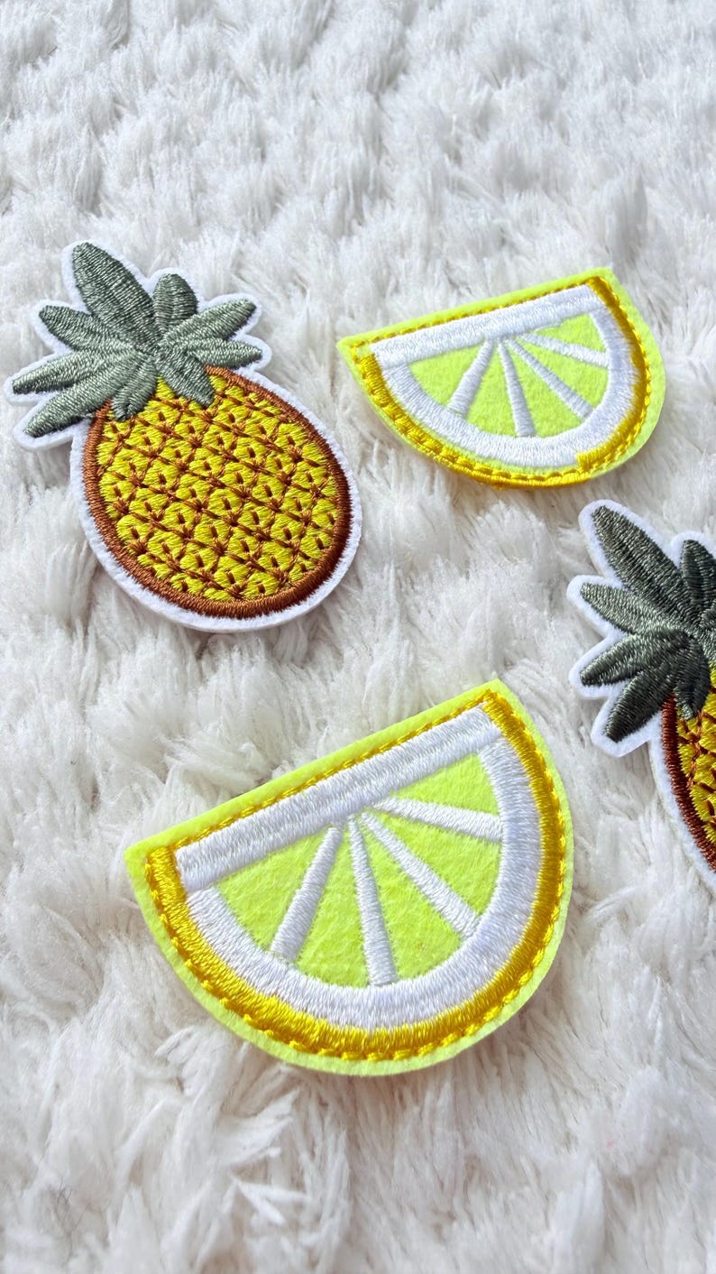 May include: Embroidered patches of a pineapple and lemon slices. The pineapple patch is yellow with brown and green detailing. The lemon slices are yellow with white and yellow accents. The patches are on a white, textured background.