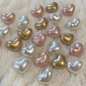 May include: A collection of heart-shaped beads in various colors, including white, pink, and gold. The beads are arranged on a white fuzzy surface.