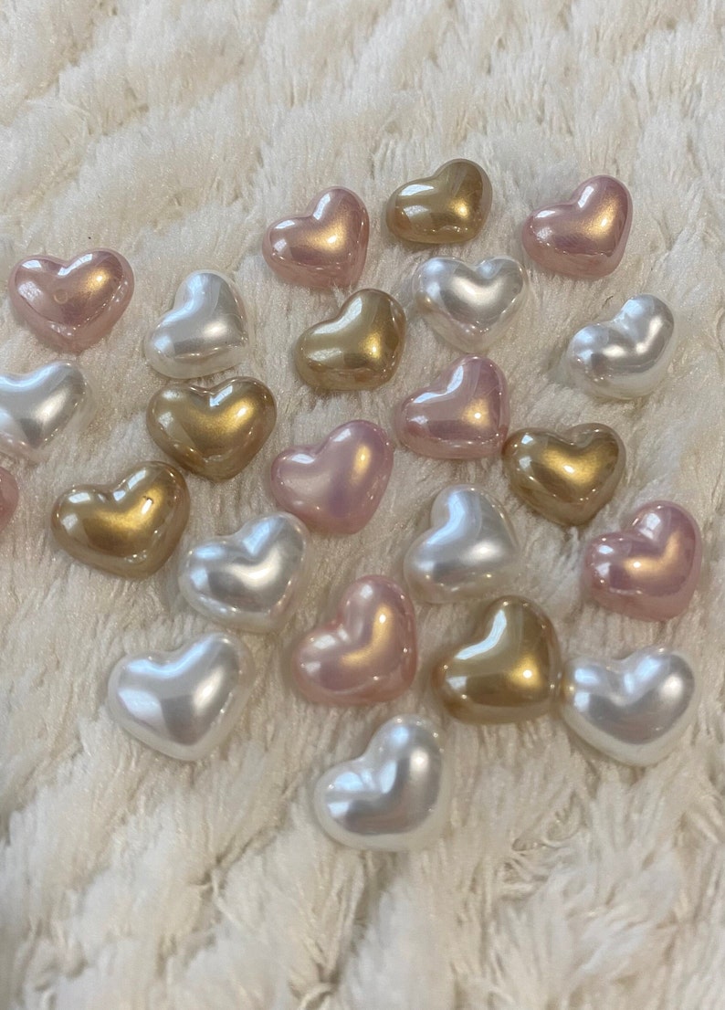 May include: A collection of heart-shaped beads in various colors, including white, pink, and gold. The beads are arranged on a white fuzzy surface.